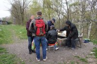 3 bezdomni za garażami , dwóch siedzi na krzesłach jeden stoi. Policjantka wita się z jednym z nich.