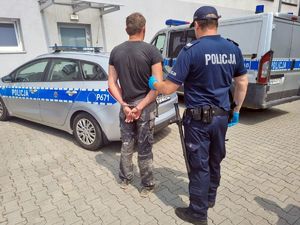 Umundurowany policjant z zatrzymanym 37-latkiem. W tle oznakowane radiowozy.