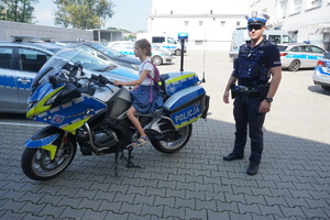 Policjant z wydziału ruchu drogowego stojący obok policyjnego motocykla, na którym siedzi dziewczynka z Placówki Wsparcia Dziennego w Niewiadomiu.