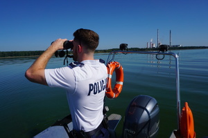 Umundurowany policjant patrzący przez lornetki na linie brzegową dba o bezpieczeństwo osób wypoczywających nad wodą.