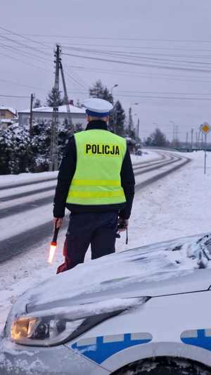 Policjant stoi przy drodze przed radiowozem. Trzyma w ręku latarkę i obserwuje ulice.