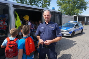 Na zdjęciu umundurowany policjant, który oprowadza dzieci. W tle radiowóz oraz dzieci.