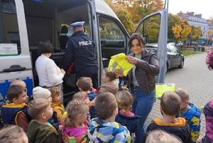 Na zdjęciu policjant rozdaje dzieciom kamizelki odblaskowe oraz opowiada o bezpiecznych zrachowaniach na drodze.