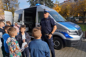 Na zdjęciu policjant rozdaje dzieciom kamizelki odblaskowe oraz opowiada o bezpiecznych zrachowaniach na drodze.