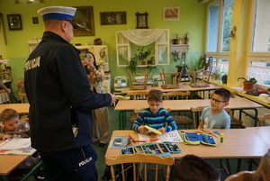 Na zdjęciu umundurowany policjant ruchu drogowego rozdaje dzieciom kamizelki odblaskowe.