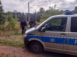 Na zdjęciu oznakowany radiowóz, a w tle policjanci.